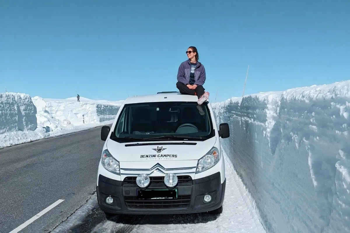 Girl traveling by campervan in April sits on its roof between snow walls in Jotunheimen NP in Norway